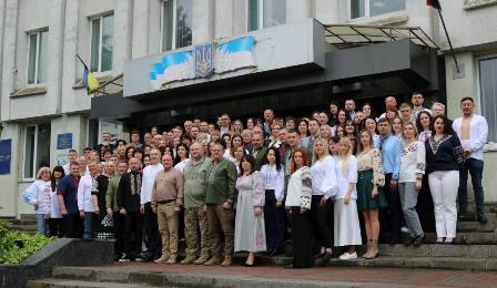 Велика група людей у вишиванках стоїть на сходах перед адміністративною будівлею з гербом України та прапорами. Колективне фото до Дня вишиванки."
