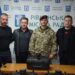 alt="На фото — група з чотирьох чоловіків на фоні банера з написом "Рівненська міська рада". Троє з них одягнені в цивільний одяг, один — у військовій формі з береткою. Перед ними на столі розташовані сучасні дрони у захисних кейсах — ймовірно, передача техніки для потреб Збройних Сил України."