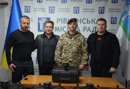 alt="На фото — група з чотирьох чоловіків на фоні банера з написом "Рівненська міська рада". Троє з них одягнені в цивільний одяг, один — у військовій формі з береткою. Перед ними на столі розташовані сучасні дрони у захисних кейсах — ймовірно, передача техніки для потреб Збройних Сил України."