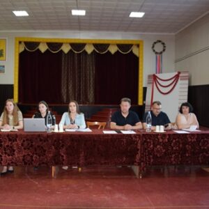 alt="Робоча зустріч у залі будинку культури: шестеро людей сидять за столом, вкритим бордовою скатертиною, перед сценою з завісами; на столі – документи, ноутбук, вода."