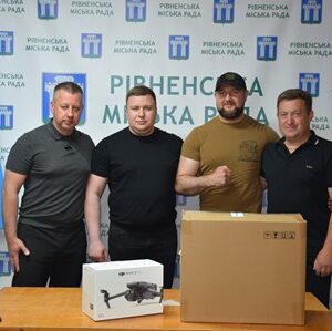 alt="Чотири чоловіки стоять поруч за столом із коробками дронів на тлі банера «Рівненська міська рада». На столі видно упаковку від дрона DJI. Поруч встановлені прапори України та Рівного."