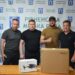 alt="Чотири чоловіки стоять поруч за столом із коробками дронів на тлі банера «Рівненська міська рада». На столі видно упаковку від дрона DJI. Поруч встановлені прапори України та Рівного."