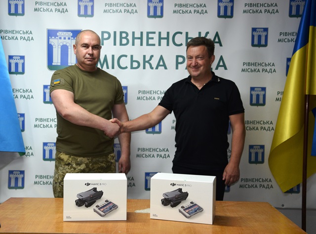 Alt="На знімку зображено урочисте передавання дронів військовим. Двоє чоловіків — військовослужбовець у формі та посадовець у цивільному одязі — тиснуть один одному руки на фоні брендового банера Рівненської міської ради. Перед ними на столі розміщено дві коробки з квадрокоптерами DJI Mavic 3 Pro. "