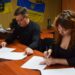 alt="Чоловік і жінка сидять за столом в офісі та підписують офіційні документи. На фоні — державний прапор України та прапор військового підрозділу. "