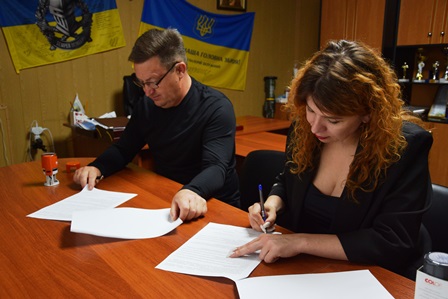alt="Чоловік і жінка сидять за столом в офісі та підписують офіційні документи. На фоні — державний прапор України та прапор військового підрозділу. "