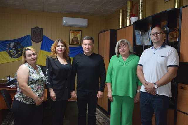 alt="На фото п’ятеро людей стоять у службовому кабінеті на фоні державної символіки України — прапора, герба та ікони."