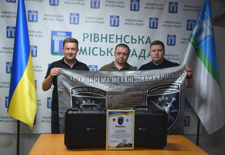 alt="Троє чоловіків тримають банер 42-ї окремої механізованої бригади ЗСУ на фоні банеру Рівненської міської ради. Перед ними на столі — два кейси з обладнанням і подяка від військових. "