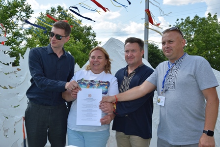 alt="Четверо людей (двоє чоловіків і одна жінка у вишиванках, один чоловік у темній сорочці та сонцезахисних окулярах) тримають у руках підписаний меморандум. Фото зроблено на відкритому повітрі під час фестивалю — на тлі білих декоративних елементів та кольорових стрічок, що майорять на вітрі. Атмосфера урочиста та позитивна. Подія пов’язана з підписанням меморандуму щодо започаткування щорічного фольклорного фестивалю «Вишнева гора»."