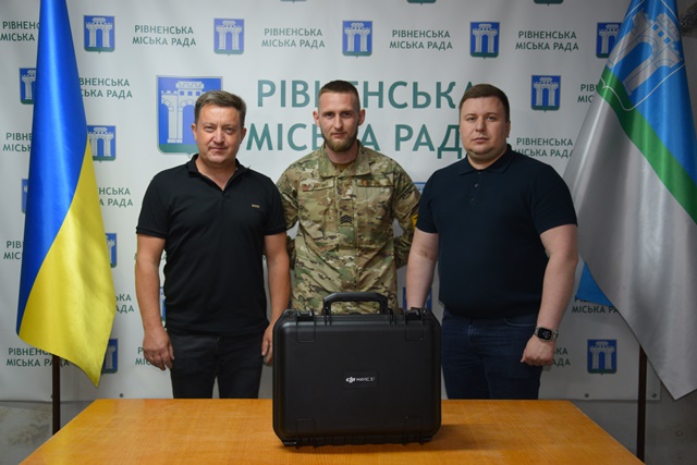 alt="Троє чоловіків, серед яких військовослужбовець у формі, стоять перед банером Рівненської міської ради. Перед ними на столі — один захисний кейс з обладнанням. "