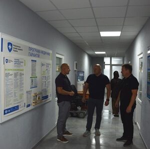 alt="Троє чоловіків ведуть розмову в коридорі адміністративної чи медичної установи. На стінах розміщені інформаційні стенди з роз’ясненнями щодо медичних гарантій, програм і послуг. "