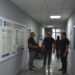alt="Троє чоловіків ведуть розмову в коридорі адміністративної чи медичної установи. На стінах розміщені інформаційні стенди з роз’ясненнями щодо медичних гарантій, програм і послуг. "