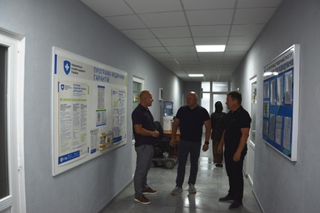alt="Троє чоловіків ведуть розмову в коридорі адміністративної чи медичної установи. На стінах розміщені інформаційні стенди з роз’ясненнями щодо медичних гарантій, програм і послуг. "