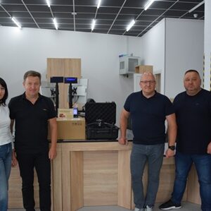 alt="група людей стоїть у приміщенні відділення поштового оператора. Перед ними — комплекти техніки у транспортних кейсах та коробках, серед яких видно дрон. Усміхнені люди — ймовірно, представники громади та працівники відділення."
