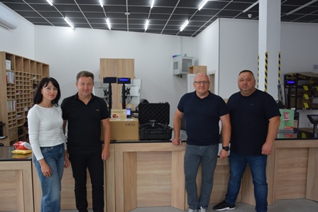 alt="група людей стоїть у приміщенні відділення поштового оператора. Перед ними — комплекти техніки у транспортних кейсах та коробках, серед яких видно дрон. Усміхнені люди — ймовірно, представники громади та працівники відділення."