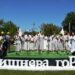 alt="На фото — сцена просто неба, оформлена декоративним написом «Вишнева гора». На сцені виступає фольклорний колектив у традиційному білому одязі, дехто тримає гілочки зелені. Поруч — музиканти з баяном і народними інструментами. На тлі — зелені дерева, прикрашені стрічками, синє небо та літня атмосфера. Фото зроблено під час першого фольклорного фестивалю «Вишнева гора» в селі Городок."