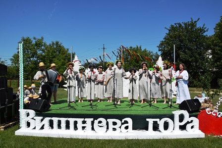 alt="На фото — сцена просто неба, оформлена декоративним написом «Вишнева гора». На сцені виступає фольклорний колектив у традиційному білому одязі, дехто тримає гілочки зелені. Поруч — музиканти з баяном і народними інструментами. На тлі — зелені дерева, прикрашені стрічками, синє небо та літня атмосфера. Фото зроблено під час першого фольклорного фестивалю «Вишнева гора» в селі Городок."