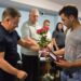 alt="Чоловік у темній футболці передає квіти та нагороду іншому чоловікові у сірій футболці. Церемонія відбувається в офіційному приміщенні, на тлі присутні люди, що стоять, вшановуючи момент. Фото зроблено під час урочистого вручення державної нагороди родичу загиблого Захисника України. Атмосфера зворушлива та офіційна."