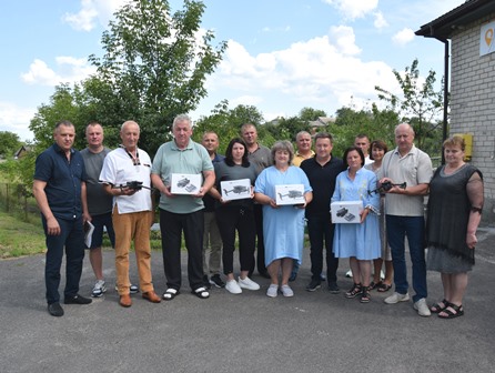 alt="Група представників Городоцької громади під час передачі квадрокоптерів для Збройних Сил України. Люди тримають у руках коробки з дронами та самі квадрокоптери. Знімок зроблений на вулиці біля адміністративної будівлі, на тлі дерев та ясного неба."