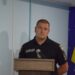alt="Чоловік у чорній формі з написом «Поліція» виступає за трибуною перед мікрофоном. На фоні — синій інформаційний банер із написом «Городоцька сільська рада» та державний прапор України."