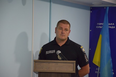 alt="Чоловік у чорній формі з написом «Поліція» виступає за трибуною перед мікрофоном. На фоні — синій інформаційний банер із написом «Городоцька сільська рада» та державний прапор України."