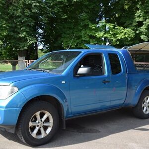 alt="На зображенні — синій пікап Nissan Navara, припаркований на асфальтованій ділянці біля зеленої зони з деревами. Автомобіль має подвійну кабіну та тентоване багажне відділення з відкритим боковим вікном. На тлі видно частину огорожі та парк."