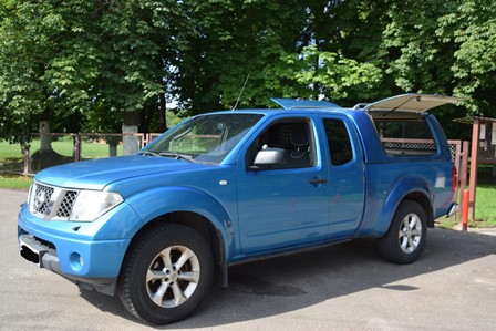 alt="На зображенні — синій пікап Nissan Navara, припаркований на асфальтованій ділянці біля зеленої зони з деревами. Автомобіль має подвійну кабіну та тентоване багажне відділення з відкритим боковим вікном. На тлі видно частину огорожі та парк."