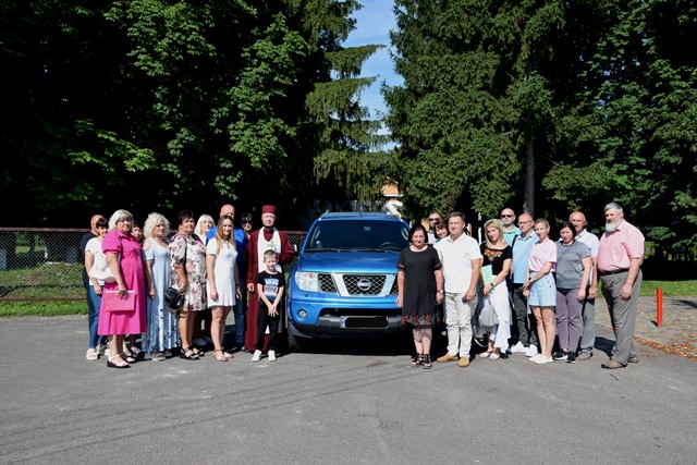 alt="Групове фото людей перед синім пікапом Nissan Navara, припаркованим на асфальтованій території на фоні зеленої паркової зони. У центрі кадру — автомобіль, з обох боків якого стоять дорослі та діти. Серед присутніх — священник, представники громади, жінки та чоловіки різного віку. Атмосфера події урочиста й доброзичлива."