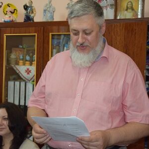 alt="Чоловік із сивою бородою у рожевій сорочці читає документ під час офіційного заходу. На задньому плані — шафа з папками, сувенірами та іконами. Поряд сидять жінки, що уважно слухають."