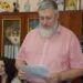 alt="Чоловік із сивою бородою у рожевій сорочці читає документ під час офіційного заходу. На задньому плані — шафа з папками, сувенірами та іконами. Поряд сидять жінки, що уважно слухають."