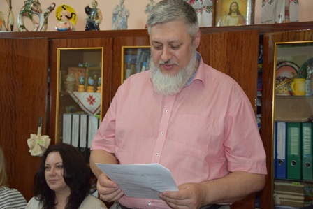 alt="Чоловік із сивою бородою у рожевій сорочці читає документ під час офіційного заходу. На задньому плані — шафа з папками, сувенірами та іконами. Поряд сидять жінки, що уважно слухають."