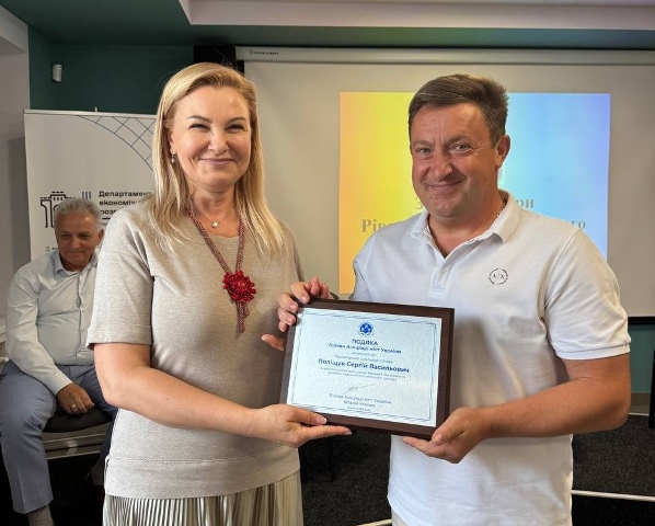 alt="жінка та чоловік тримають у руках подяку від Голови Асоціації міст України. Чоловік — Городоцький сільський голова Сергій Поліщук — отримує відзнаку за допомогу Збройним Силам України та розвиток громади. Подія відбувається у приміщенні, на фоні видно банер та екран із презентацією."