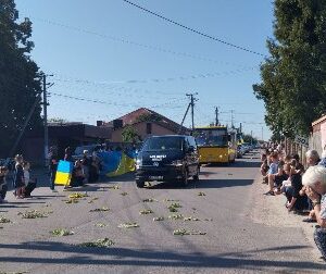 Прощальна церемонія у селі: люди стоять і сидять на колінах обабіч дороги, вшановуючи загиблого Захисника. Посеред вулиці — катафалк, прикрашений квітами, попереду — державний прапор України, люди з прапорами та квітами. На дорозі розкидані зелені гілки. Атмосфера скорботи та поваги
