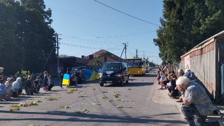 Прощальна церемонія у селі: люди стоять і сидять на колінах обабіч дороги, вшановуючи загиблого Захисника. Посеред вулиці — катафалк, прикрашений квітами, попереду — державний прапор України, люди з прапорами та квітами. На дорозі розкидані зелені гілки. Атмосфера скорботи та поваги
