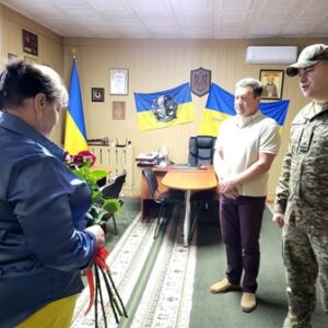 alt="У кабінеті Городоцької сільської ради триває урочиста церемонія: мати загиблого воїна тримає букет червоних троянд, перед нею — очільник громади Сергій Поліщук та військовослужбовець Національної гвардії України. На стіні — державні та військові прапори, портрети й герб України."