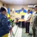 alt="У кабінеті Городоцької сільської ради триває урочиста церемонія: мати загиблого воїна тримає букет червоних троянд, перед нею — очільник громади Сергій Поліщук та військовослужбовець Національної гвардії України. На стіні — державні та військові прапори, портрети й герб України."