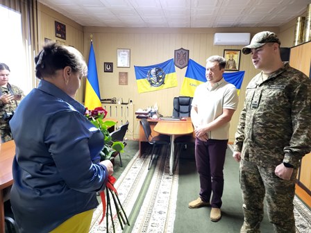 alt="У кабінеті Городоцької сільської ради триває урочиста церемонія: мати загиблого воїна тримає букет червоних троянд, перед нею — очільник громади Сергій Поліщук та військовослужбовець Національної гвардії України. На стіні — державні та військові прапори, портрети й герб України."