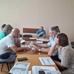 Учасники засідання сидять за дерев'яним столом, аналізуючи документи з таблицями та текстами. Один із чоловіків у блакитній футболці веде обговорення, інші уважно слухають та роблять нотатки.