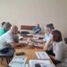 Учасники засідання сидять за дерев'яним столом, аналізуючи документи з таблицями та текстами. Один із чоловіків у блакитній футболці веде обговорення, інші уважно слухають та роблять нотатки.