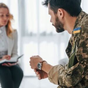Чоловік у військовій формі з нашивкою українського прапора сидить, схиливши голову та стискаючи руки. Перед ним на стільці сидить жінка в білому піджаку, яка тримає блокнот і ручку, ніби проводить консультацію або бесіду. Світла, простора кімната з великими вікнами на задньому плані.