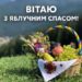 Кошик з фруктами та запашними травами, прикрашений вишитим рушником, стоїть на зеленій траві на тлі гір. У кошику — яблука, виноград, груша, а також букет із польових квітів. Надпис: “Вітаю з Яблучним Спасом!”