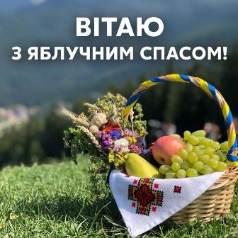 Кошик з фруктами та запашними травами, прикрашений вишитим рушником, стоїть на зеленій траві на тлі гір. У кошику — яблука, виноград, груша, а також букет із польових квітів. Надпис: “Вітаю з Яблучним Спасом!”