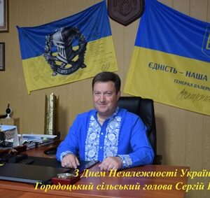 Чоловік у вишиванці сидить за робочим столом у кабінеті на тлі українських прапорів. Текст на фото: 'З Днем Незалежності України! Городоцький сільський голова Сергій Поліщук