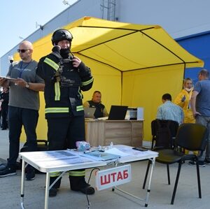 На вулиці біля будівлі розгорнуто жовтий намет із табличкою «ШТАБ». Перед наметом стоїть рятувальник у повному захисному спорядженні та касці, поруч — чоловік із блокнотом. У наметі працюють люди, а на задньому плані видно учасників у жовтих захисних костюмах. Ймовірно, проходять навчання або тренування з надзвичайних ситуацій.