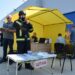 На вулиці біля будівлі розгорнуто жовтий намет із табличкою «ШТАБ». Перед наметом стоїть рятувальник у повному захисному спорядженні та касці, поруч — чоловік із блокнотом. У наметі працюють люди, а на задньому плані видно учасників у жовтих захисних костюмах. Ймовірно, проходять навчання або тренування з надзвичайних ситуацій.
