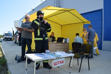 На вулиці біля будівлі розгорнуто жовтий намет із табличкою «ШТАБ». Перед наметом стоїть рятувальник у повному захисному спорядженні та касці, поруч — чоловік із блокнотом. У наметі працюють люди, а на задньому плані видно учасників у жовтих захисних костюмах. Ймовірно, проходять навчання або тренування з надзвичайних ситуацій.