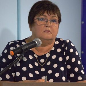 Жінка у темній сукні в білий горох виступає на трибуні з мікрофоном під час сесії сільської ради.