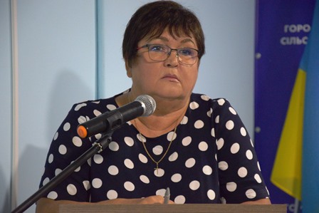 Жінка у темній сукні в білий горох виступає на трибуні з мікрофоном під час сесії сільської ради.