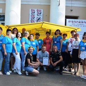 Групове фото учасників заходу біля жовтого намету з написом “Гориньські…”. Люди одягнені у сині футболки з жовтими хустками та інші святкові вбрання, на задньому плані — будівля з колонами та виставкові банери