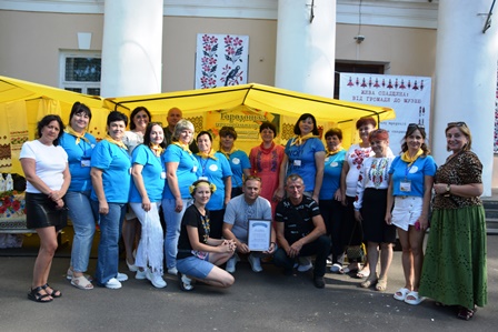 Групове фото учасників заходу біля жовтого намету з написом “Гориньські…”. Люди одягнені у сині футболки з жовтими хустками та інші святкові вбрання, на задньому плані — будівля з колонами та виставкові банери