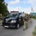 Чорний мікроавтобус із написом «НА СЛУЖБІ ЖИТТЯ» рухається сільською дорогою, поруч із ним крокують військовослужбовці у формі та зі зброєю. На задньому плані видно приватні будинки та зелені насадження
